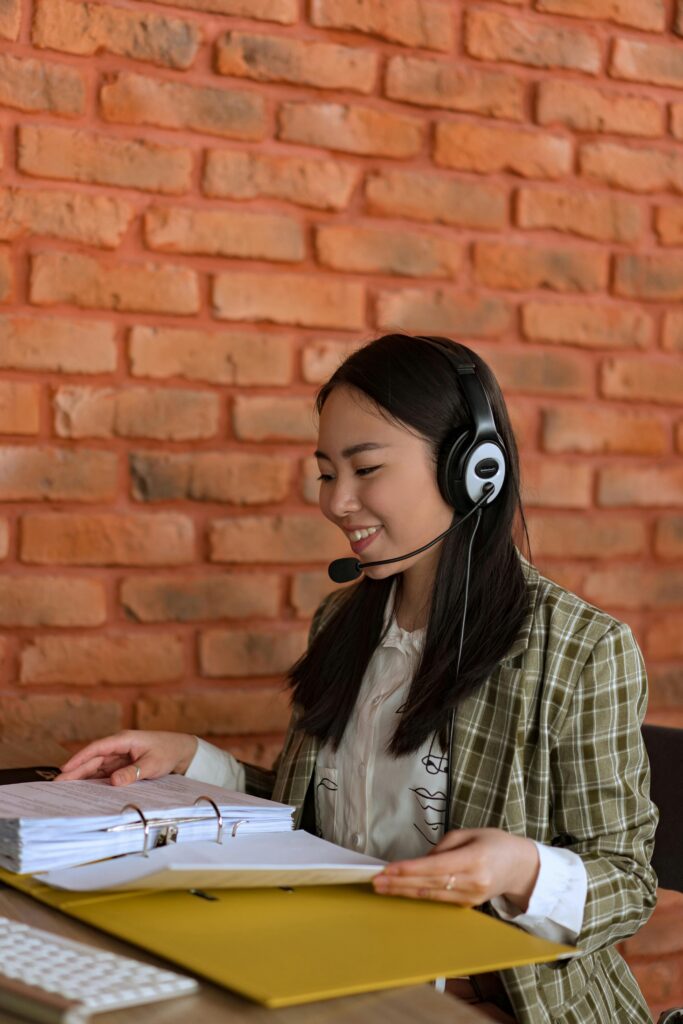 pexels photo 8101449 8101449 Professional Asian woman wearing a headset, working efficiently at an office with a binder, smiling and focused.